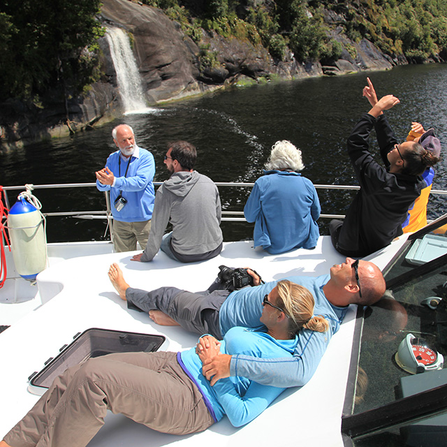 Guests relaxing on the bow of the Southern Secret and looking high up to one of the many waterfalls of Doubtful Sound.